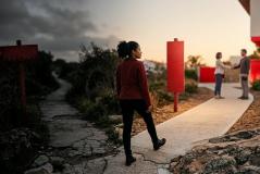 Pedestre de casaco vermelho e cabelos cacheados decide entre dois caminhos: uma trilha de terra escura e danificada sob um céu nublado, e uma calçada clara e iluminada pelo sol que guia em direção a pessoas se cumprimentando amigavelmente diante de uma casa moderna.