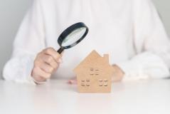 Uma mão segura uma lupa apontada para uma pequena miniatura de casa feita de papelão ou madeira clara.