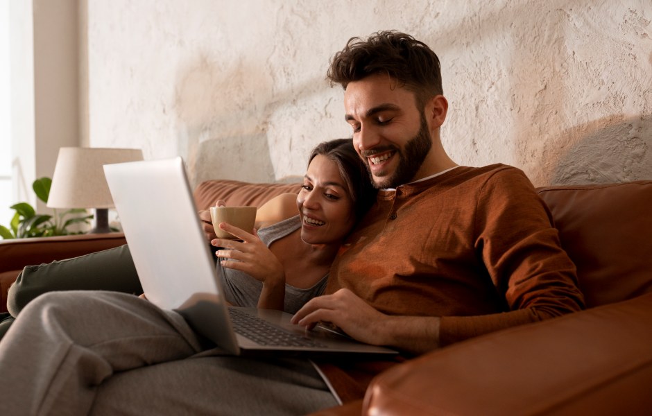 Um casal sorridente relaxa em um sofá de couro enquanto usa um notebook e toma café em um ambiente iluminado.