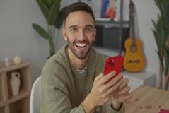 Homem sorridente segurando um smartphone vermelho em ambiente interno, com guitarra e plantas ao fundo, promovendo a palavra-chave 