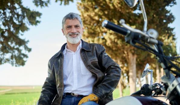 Homem sorridente posando ao lado de uma motocicleta em um parque com árvores ao fundo, usando jaqueta de couro e camisa branca.