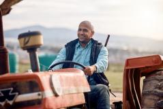 Agricultor sorridente conduz um trator vermelho no campo, com paisagem rural e montanhas ao fundo, transmitindo satisfação e orgulho no trabalho no campo.