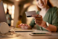 Mulher sorridente segura cartão de crédito enquanto realiza uma compra online no laptop, num ambiente com iluminação aconchegante e quente.