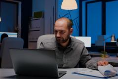 Profissional analisa relatórios e gráficos no laptop durante a madrugada em escritório com iluminação azulada, transmitindo dedicação intensa ao trabalho.
