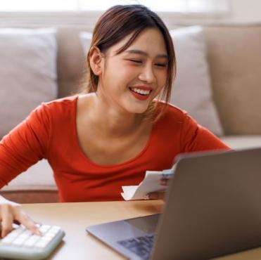 Mulher sorridente usando um laptop e segurando uma fatura, estudando ou realizando tarefas administrativas em casa, com expressão de satisfação.
