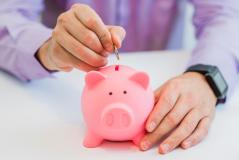 Mão depositando uma moeda em um cofrinho rosa, simbolizando o hábito de poupar e a importância do planejamento financeiro para o futuro.
