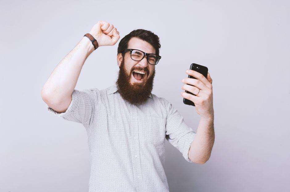  Um homem barbudo de óculos grita de alegria e comemora com o punho erguido ao olhar para o celular.