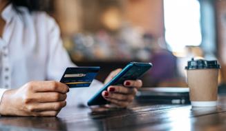 Mulher realiza pagamento pelo smartphone segurando cartão de crédito numa cafeteria, com copo de café descartável ao lado sobre a mesa.