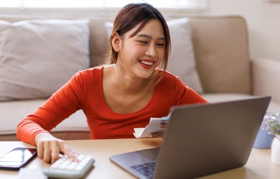 Mulher sorridente usando um laptop e segurando uma fatura, estudando ou realizando tarefas administrativas em casa, com expressão de satisfação.
