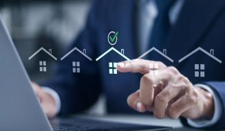 Pessoa de terno interagindo com uma tela digital que mostra ícones de casas e uma marca de verificação, simbolizando aprovação ou compra de imóveis, em um ambiente de escritório.