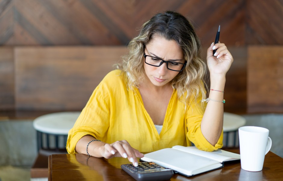 Mulher de óculos analisa contas com expressão preocupada, usando calculadora e caderno sobre a mesa, com uma xícara de café ao lado.
