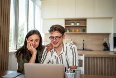 Casal sorridente assistindo a algo no laptop sentado na cozinha moderna com iluminação natural, elementos decorativos e ambiente acolhedor.