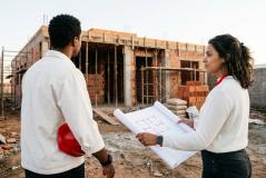 Um homem segurando um capacete de proteção vermelho e uma mulher segurando uma planta de arquitetura desenrolada, analisam juntos uma obra em um canteiro de construção com estrutura de tijolos ao fundo.