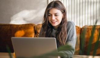 Jovem sorridente usa o laptop confortavelmente na sala de casa, com sofá mostarda e luz natural ao fundo, transmitindo bem-estar e praticidade no dia a dia.