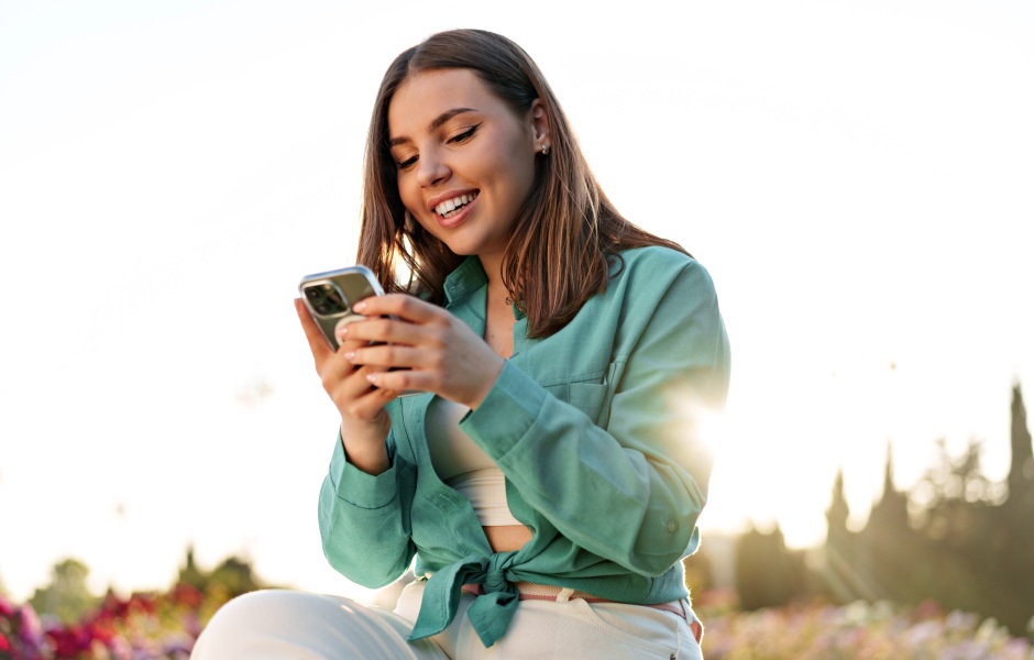 Jovem sorridente usa o smartphone ao ar livre num parque florido, com luz dourada do pôr do sol ao fundo criando uma atmosfera leve e descontraída.