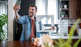 Homem feliz tirando selfie na cozinha moderna enquanto segura uma casa de papelão com uma decoração aconchegante e plantas ao redor.