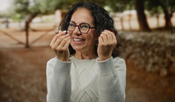 Mulher sorridente com óculos, vestindo suéter cinza, ao ar livre, expressando felicidade e alegria com um fundo de árvores e natureza.