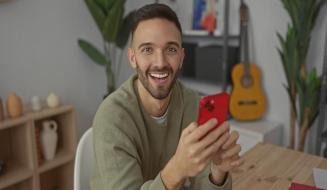 Um homem jovem e sorridente segura um celular vermelho enquanto está sentado à frente de uma mesa de madeira.