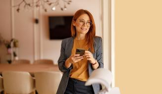Mulher com cabelo vermelho, usando óculos e roupa profissional cinza blazer e blusa na cor mostarda, sorrindo e segurando um smartphone em ambiente moderno com decoração elegante