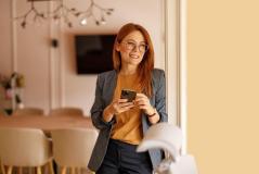 Uma mulher ruiva de óculos e blazer sorri enquanto segura um celular, encostada em um batente de porta.