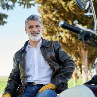 Um homem maduro de barba grisalha e jaqueta de couro sorri sentado em sua motocicleta em uma estrada arborizada.