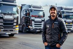 Caminhoneiro sorridente posa com jaqueta Scania em frente a uma frota de caminhões da marca, transmitindo orgulho e identificação com a profissão.