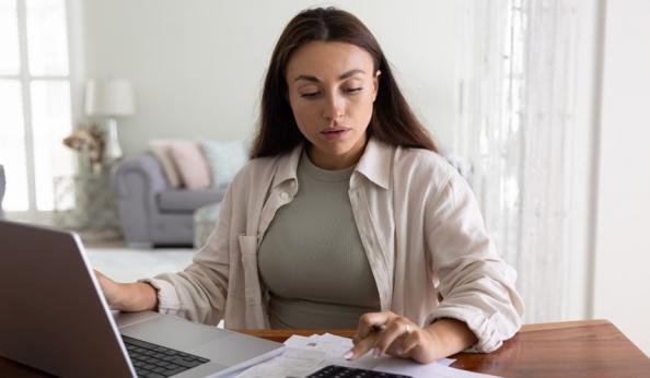 Mulher estudando contas financeiras em casa, com laptop, calculadora e papéis sobre a mesa, em um ambiente doméstico bem iluminado, ideal para gerenciamento financeiro.