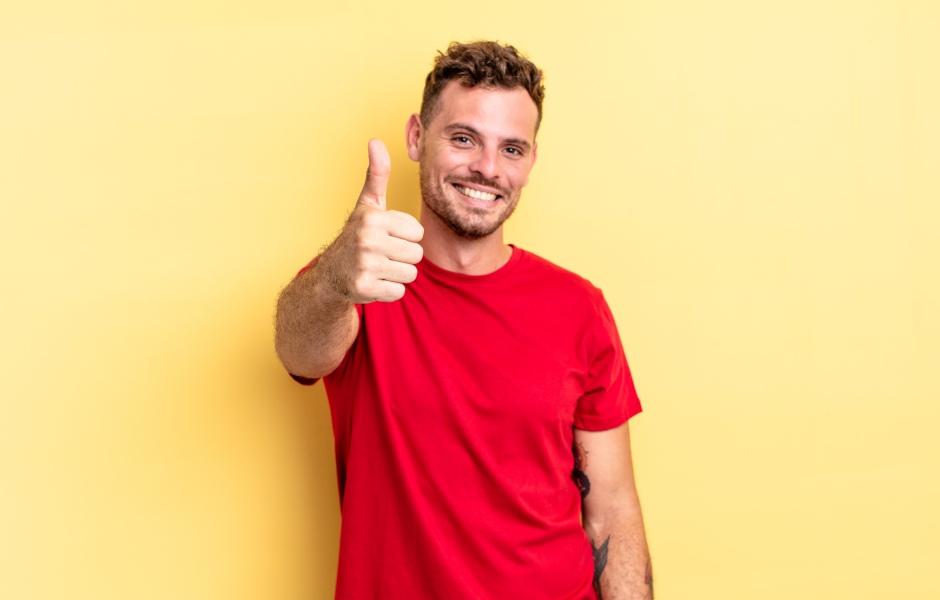 Homem jovem sorrindo com expressão positiva, fazendo sinal de joinha (polegar para cima) com a mão esquerda, vestindo camiseta vermelha, sobre fundo amarelo claro.