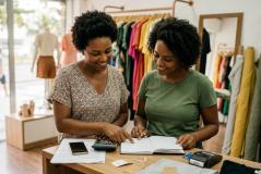 Duas empreendedoras sorriem enquanto fazem cálculos e anotações no balcão de uma loja de roupas colorida, transmitindo parceria e gestão ativa do negócio.