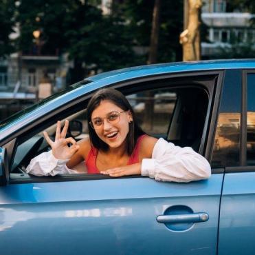 Mulher sorridente dentro de um carro azul, olhando pela janela e fazendo o sinal de 'ok' com a mão.