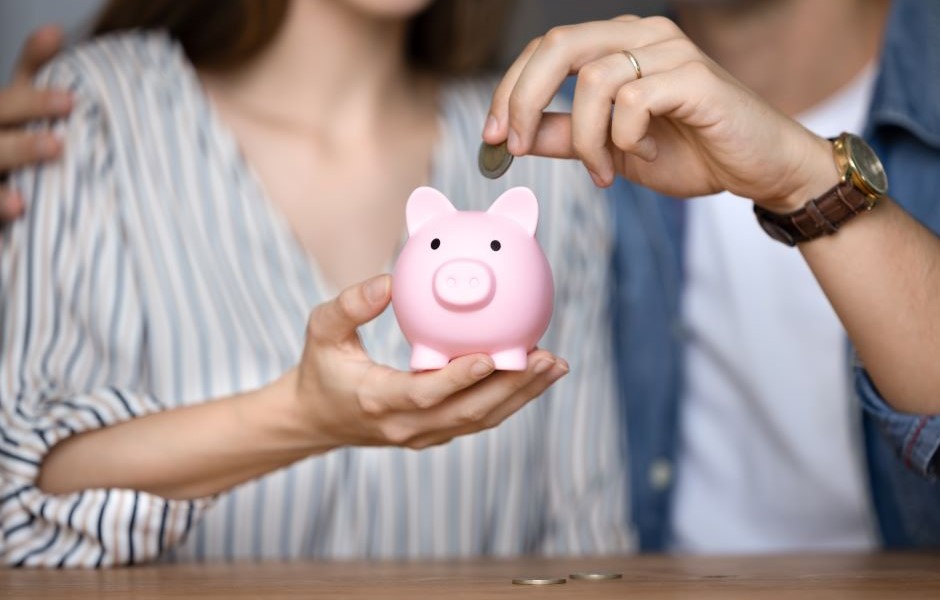 Imagem de uma pessoa colocando moedas na pequena piggy bank de cor rosa, simbolizando economia e finanças.