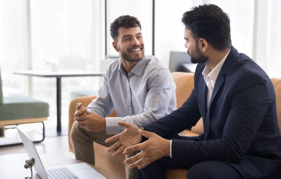Dois profissionais em trajes sociais conversam de forma amigável em um escritório moderno, com um notebook sobre a mesa.
