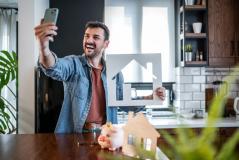 Homem feliz tirando selfie na cozinha moderna enquanto segura uma casa de papelão com uma decoração aconchegante e plantas ao redor.