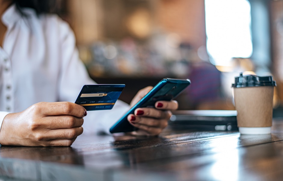 Mulher realiza pagamento pelo smartphone segurando cartão de crédito numa cafeteria, com copo de café descartável ao lado sobre a mesa.