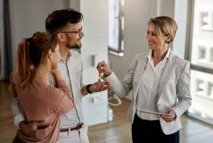 Corretora sorridente entrega as chaves do imóvel a um casal abraçado, num apartamento amplo e iluminado, celebrando a conquista da casa própria.