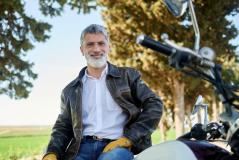Um homem maduro de barba grisalha e jaqueta de couro sorri sentado em sua motocicleta em uma estrada arborizada.