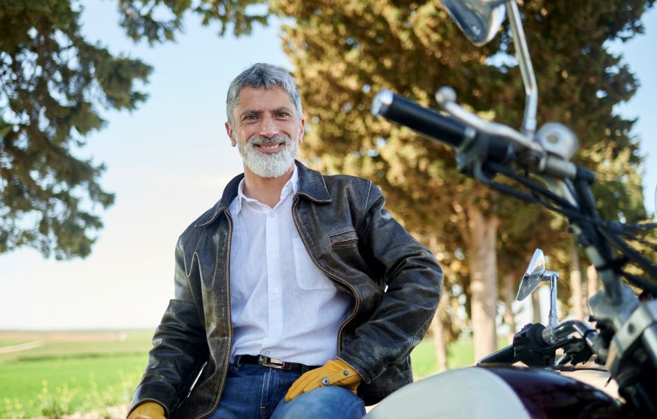 Homem sorridente posando ao lado de uma motocicleta em um parque com árvores ao fundo, usando jaqueta de couro e camisa branca.