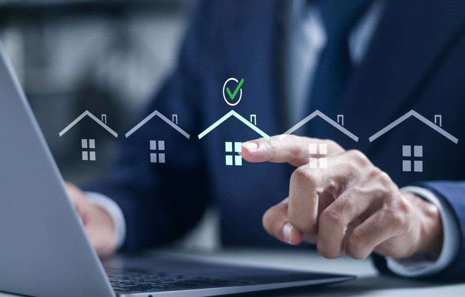 Pessoa de terno interagindo com uma tela digital que mostra ícones de casas e uma marca de verificação, simbolizando aprovação ou compra de imóveis, em um ambiente de escritório.