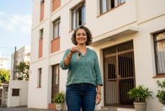 Mulher de cabelos cacheados grisalhos, camisa verde e calça jeans sorri ao segurar molho de chaves em frente a edifício residencial. Ao fundo, destaca-se fachada clara do prédio com detalhes alaranjados, janelas, portão de ferro e vasos de plantas junto à entrada.