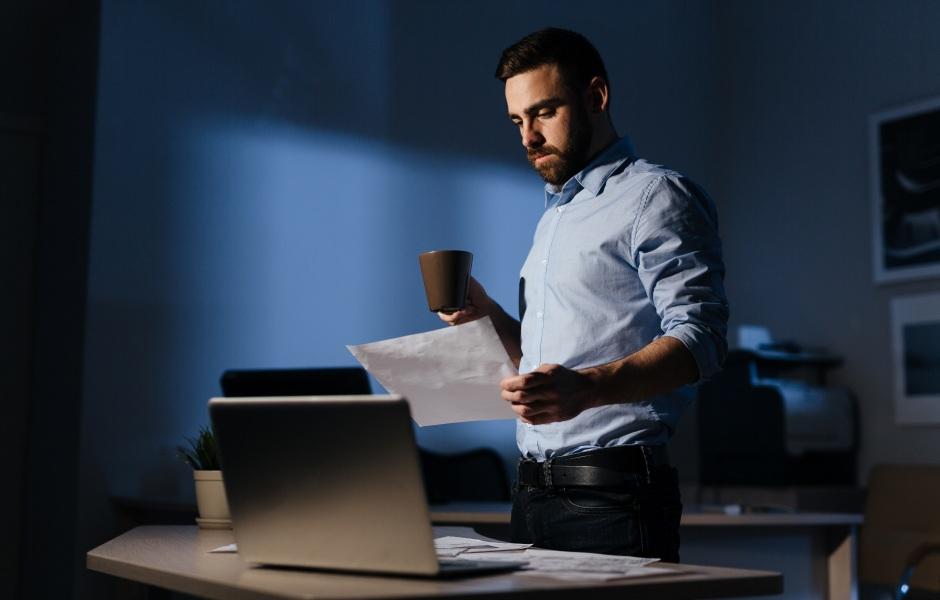 Um homem de barba veste camisa social e analisa documentos em pé, segurando uma caneca de café. Ele está em um escritório escurecido à noite, com um notebook aberto sobre a mesa à sua frente.