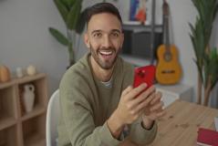 Um homem jovem e sorridente segura um celular vermelho enquanto está sentado à frente de uma mesa de madeira.
