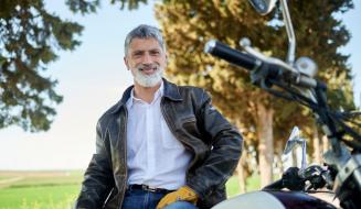 Homem sorridente posando ao lado de uma motocicleta em um parque com árvores ao fundo, usando jaqueta de couro e camisa branca.