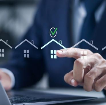 Pessoa de terno interagindo com uma tela digital que mostra ícones de casas e uma marca de verificação, simbolizando aprovação ou compra de imóveis, em um ambiente de escritório.