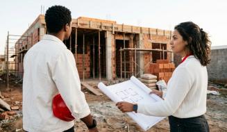 Um homem segurando um capacete de proteção vermelho e uma mulher segurando uma planta de arquitetura desenrolada, analisam juntos uma obra em um canteiro de construção com estrutura de tijolos ao fundo.