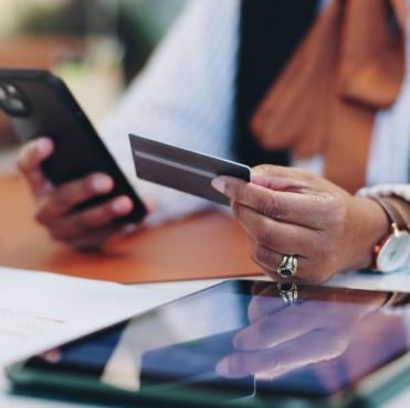 Pessoa usando um smartphone e um cartão de crédito em uma mesa de trabalho, com vários dispositivos eletrônicos ao redor, representando transações bancárias e compras online.