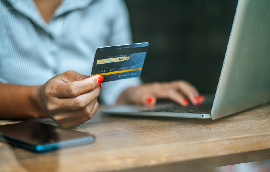 Close de mulher segurando cartão de crédito azul enquanto digita no laptop, realizando um pagamento online com celular ao lado sobre a mesa.