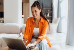 Uma mulher jovem e sorridente utiliza um notebook sobre uma mesa de centro enquanto está sentada em um sofá claro.