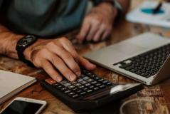 Uma mão masculina com um relógio preto utiliza uma calculadora sobre uma mesa de madeira rústica.