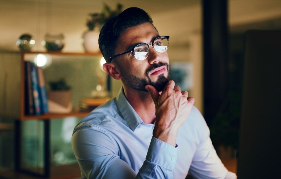 Um homem de barba e óculos olha pensativo para o lado enquanto apoia a mão no queixo em um escritório. Ele veste uma camisa social clara e está em um ambiente moderno com iluminação suave ao fundo.