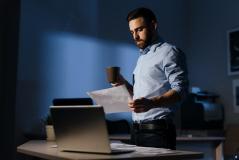 Um homem de barba veste camisa social e analisa documentos em pé, segurando uma caneca de café. Ele está em um escritório escurecido à noite, com um notebook aberto sobre a mesa à sua frente.
